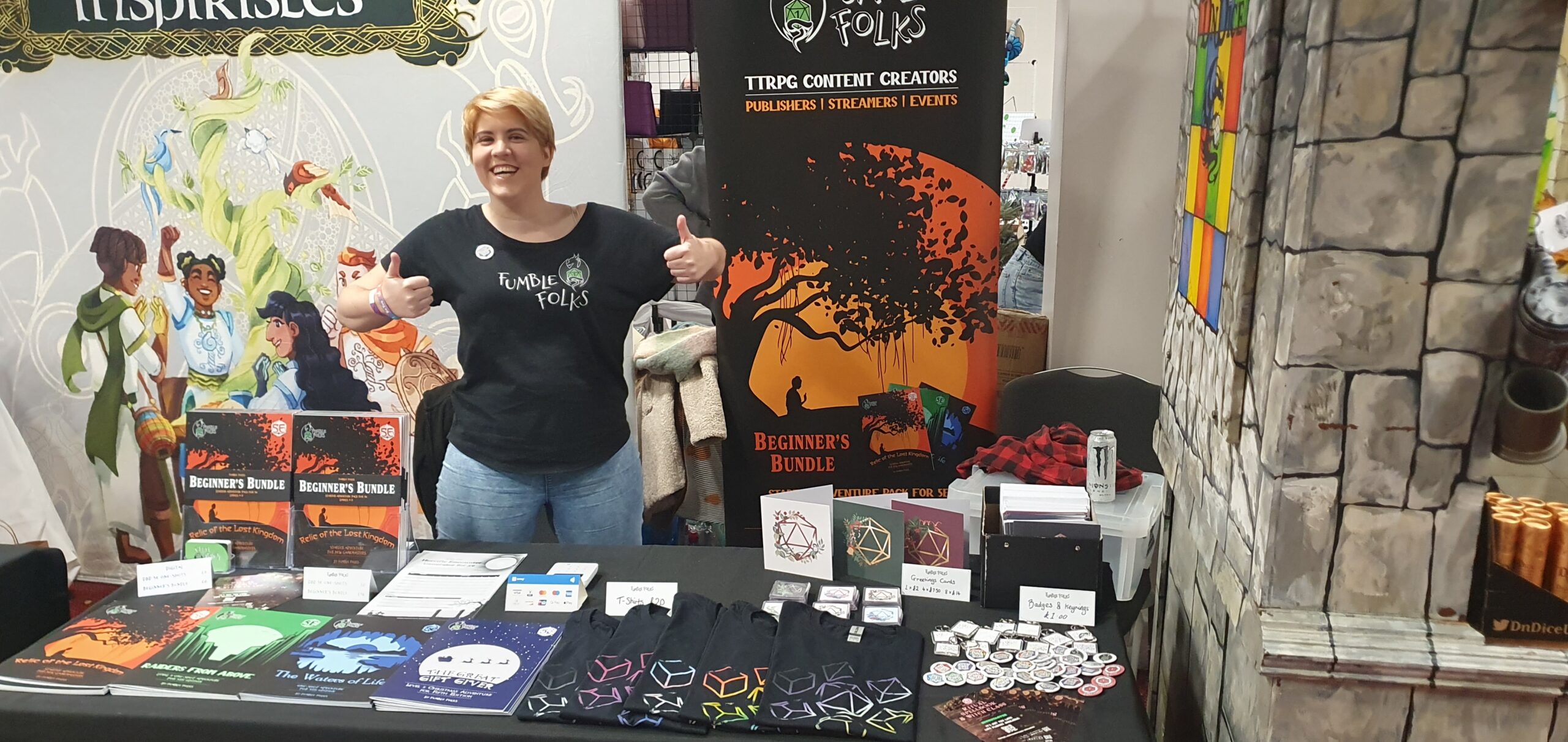 Bree, holding two thumbs up, stood behind our Dragonmeet stall, covered with our lovely merch: books, cards, tees, magnets and badges