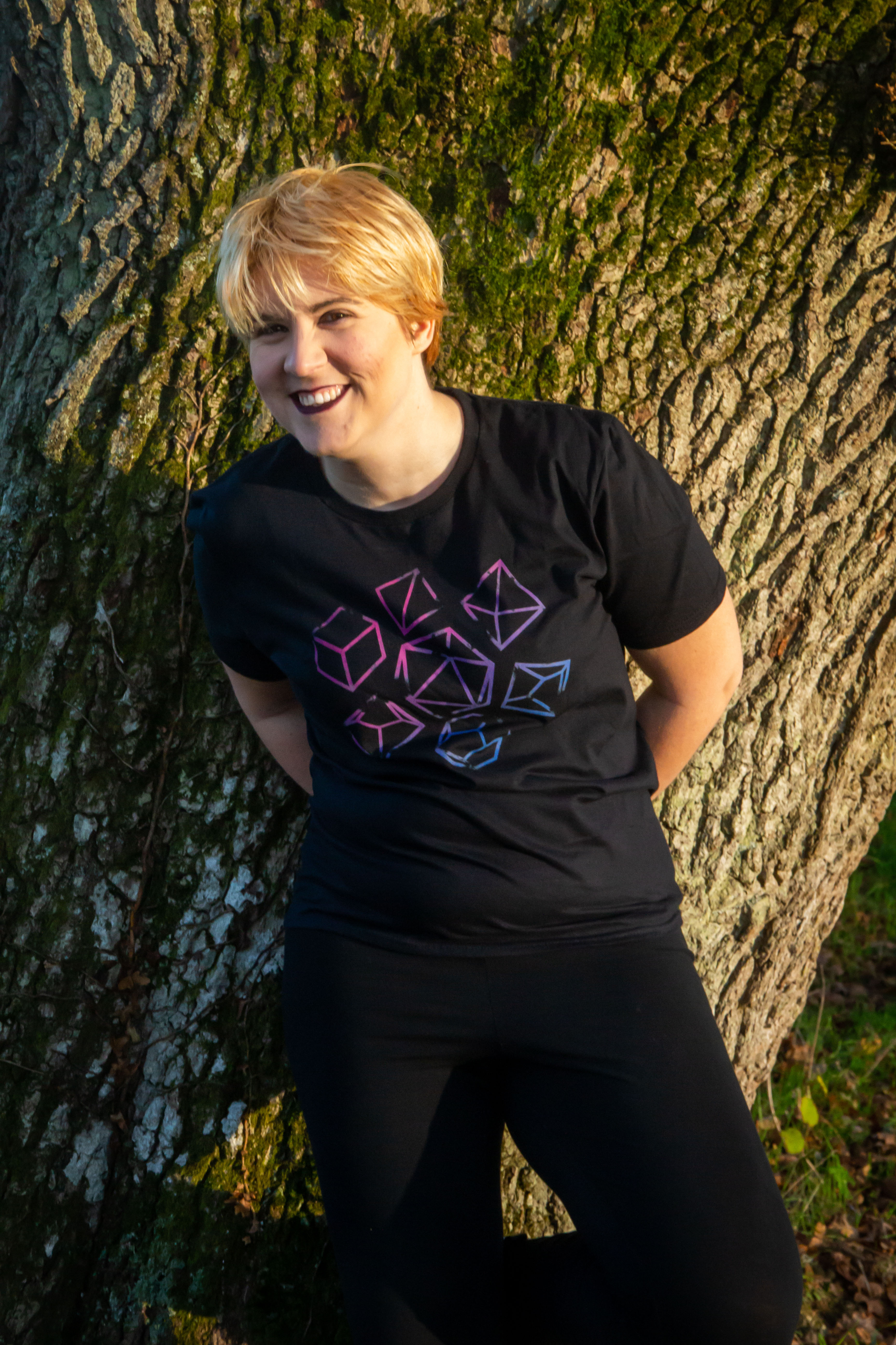 Fumble Folks Founder, Bree, pretending it's not cold, against a tree and wearing our black tee with dice pattern in bisexual colours.